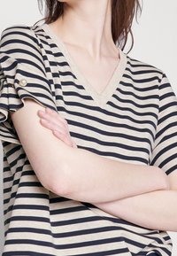 Top de cuello en V a rayas en beige y azul marino, con mangas con volantes y detalles de botones de perla. Tejido suave con rayas horizontales.