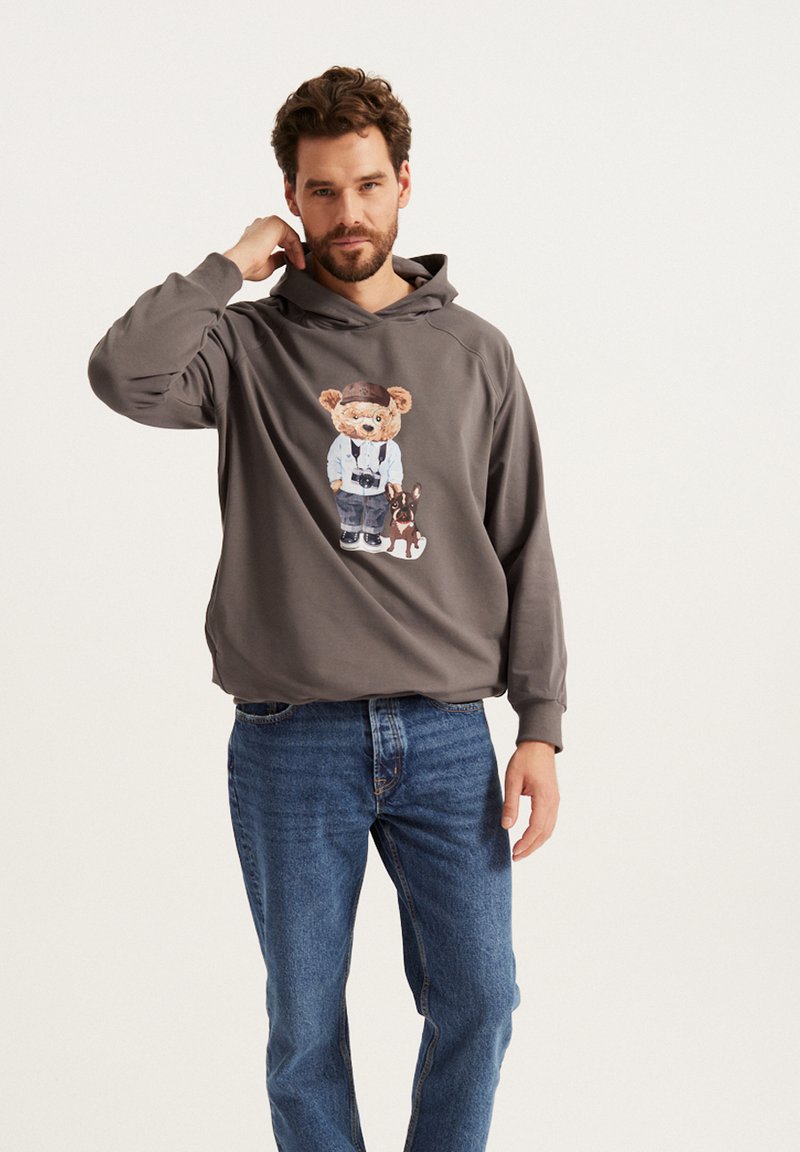 Szary hoodie z grafiką przedstawiającą niedźwiedzia i psa; miękka tkanina, luźny krój, długie rękawy i kieszeń kangurką; w zestawie z niebieskimi dżinsami.
