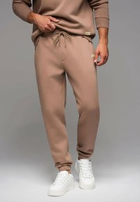 Beiga sweatpants med dragsko i midjan, ribbade muddar och små logotypdetaljer. Kombinerat med vita sneakers på en neutral bakgrund.