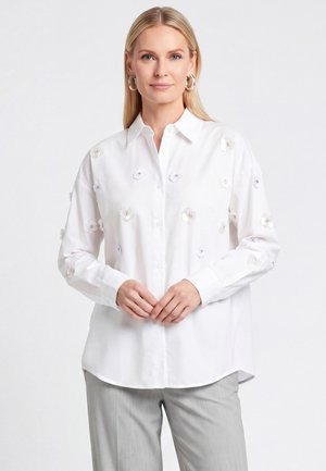 Blondynka ubrana w białą koszulę na guziki z małymi kwiatowymi zdobieniami oraz jasnoszare spodnie, stojąca z rękami złożonymi przed sobą.