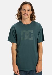 Dunkelgrünes Baumwoll-T-Shirt mit kurzen Ärmeln, das auf der Vorderseite ein großes, leicht erhabenes DC-Logo in einer helleren Grünton zeigt.