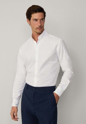 Witte button-up shirt met een spread collar, lange mouwen en contrasterende gele knopen, gedragen met donkerblauwe broeken. Eenvoudig ontwerp, geen patronen.
