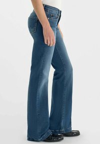 Jeans bleu évasés en denim avec des coutures visibles, une taille mi-haute et une texture légèrement usée. Portés avec des chaussures noires.