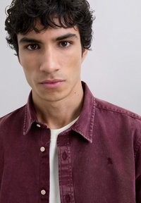 Burgundowa koszula z kołnierzykiem na guziki, miękka w dotyku, z detalem kieszonki z przodu, nałożona na białą koszulę.