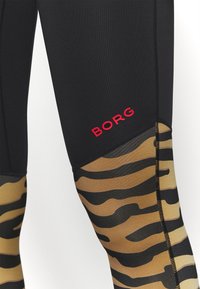 Czarne legginsy sportowe z wzorem w tygrysie paski w odcieniach beżu i czerni na dolnej części nogawki, z czerwonym logo "BORG" z boku.