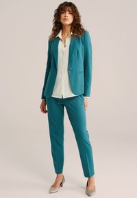 Zeleni blazer in hlače iz gladkega blaga, združeni z belo srajco s črtami. Imajo en gumb za zapiranje in konične pete.