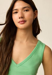Top verde senza maniche con scollo a V, realizzato in morbido cotone e con cuciture pulite, indossato da un modello con lunghi capelli scuri.