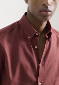 Camisa de lino roja de botón con cuello extendido, que muestra un ligero brillo y una textura sutil. Presenta botones blancos y un pequeño detalle de logo.