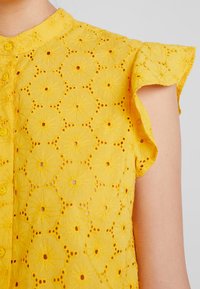 Yellow blouse featuring floral embroidery, button closure, and ruffled cap sleeves. The fabric has cut-out patterns for a textured appearance.