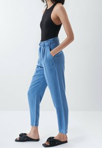 Jeans de tiro alto azul claro con un frente plisado, bolsillos laterales y un ajuste relajado, combinados con un bodysuit negro y sandalias negras de tipo slide.