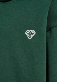 Grün gestricktes Kleidungsstück mit weicher Textur, das ein weißes, aufgesticktes Logo in Form einer Biene auf der Brust zeigt. Einfaches Design und runder Ausschnitt.