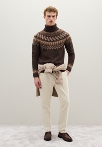 Brown knitted turtleneck sweater with geometric pattern, paired with cream corduroy pants. Brown shoes, with a light-colored sweater tied around the waist.