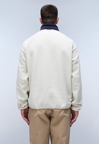 Hvid fleece jakke med en marineblå krave, der har en afslappet pasform og glat tekstur, parret med beige bukser. Vist fra bagfra.