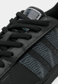 Μαύρο δερμάτινο αθλητικό παπούτσι με υφασμένους γκρι τόνους, τρεις πλαϊνές ρίγες, μαύρα κορδόνια και ετικέτα με το λογότυπο στη γλώσσα.