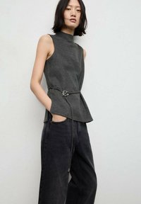 Top grigio senza maniche con collo alto e design peplum, stretto in vita con una sottile cintura con fibbia argentata, abbinato a jeans neri.