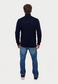 Hombre de pie mirando hacia atrás, vestido con un suéter de cuello alto azul marino, jeans azules y zapatillas negras con suelas blancas.