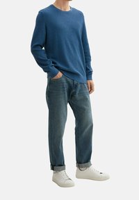 Maglione a coste blu, jeans azzurri chiari e sneakers bianche. Il maglione ha un collo rotondo e una finitura testurizzata. I jeans sono arrotolati alla caviglia.