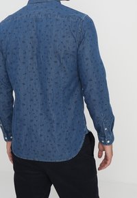 Camisa de mezclilla azul de manga larga, con puños abotonados, cuello tipo inglés y un sutil patrón floral. Hembra curva en la parte posterior.