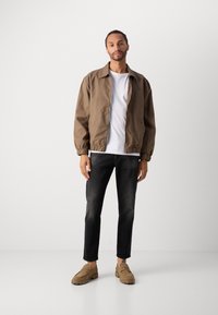 Jachetă bomber maro cu manșete elastice, tricou alb, blugi din denim închis și pantofi slip-on de culoare maro deschis, plasate pe un fundal simplu.