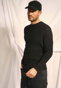 Černý pletený svetr s žebrovanou texturou, kulatým výstřihem a slim fit, kombinovaný s tmavými džíny a černou čepicí. Měkký materiál, neformální design.