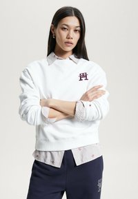 Felpa bianca con polsini e scollatura a costine, con un logo bordeaux. Indossata sopra una maglietta a righe grigio chiaro e bianco. Pantaloni scuri.