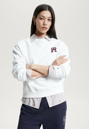 Felpa bianca con polsini e scollatura a costine, con un logo bordeaux. Indossata sopra una maglietta a righe grigio chiaro e bianco. Pantaloni scuri.
