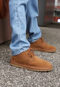 Světle modré džíny na sobě s hnědými semišovými UGG šněrovacími botami stojícími na flekaté podlaze.