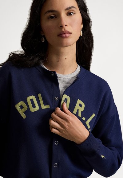 Jonge vrouw met donker haar, gekleed in een marineblauwe sweatvest met knopen en de letters "P O L O R.L.", een knoop vastmakend, neutrale achtergrond.