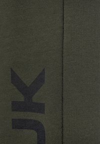 Olijfgroene stof met een gestructureerd breipatroon. Heeft een prominent zwart logo-ontwerp. Naaddetail zichtbaar aan één kant.