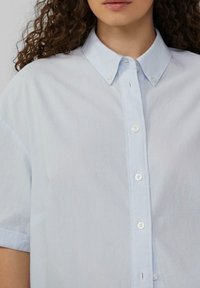 Jasnoniebieska koszula z krótkim rękawem zapinana na guziki, z kołnierzykiem i pionowymi białymi paskami. Posiada listwę z przodu z białymi guzikami i luźny krój.