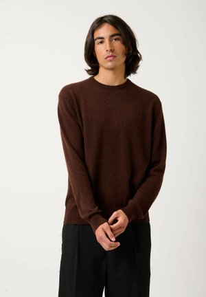 Pull en maille marron avec un col ras du cou à côtes, manches longues et une texture douce, associé à un pantalon noir. Fond blanc uni.
