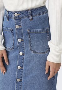 Denim szoknya elülső gombos záródással, két nagy zsebbel, világoskék színben és kontrasztos varrással. A bordázott fehér pulóver ujja látható.