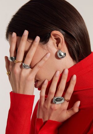 Silberohrringe mit runden Formen, großer silberner Ring, goldener gedrehter Ring und roter gerippter Rollkragenpullover. Die Nägel sind natürlich.