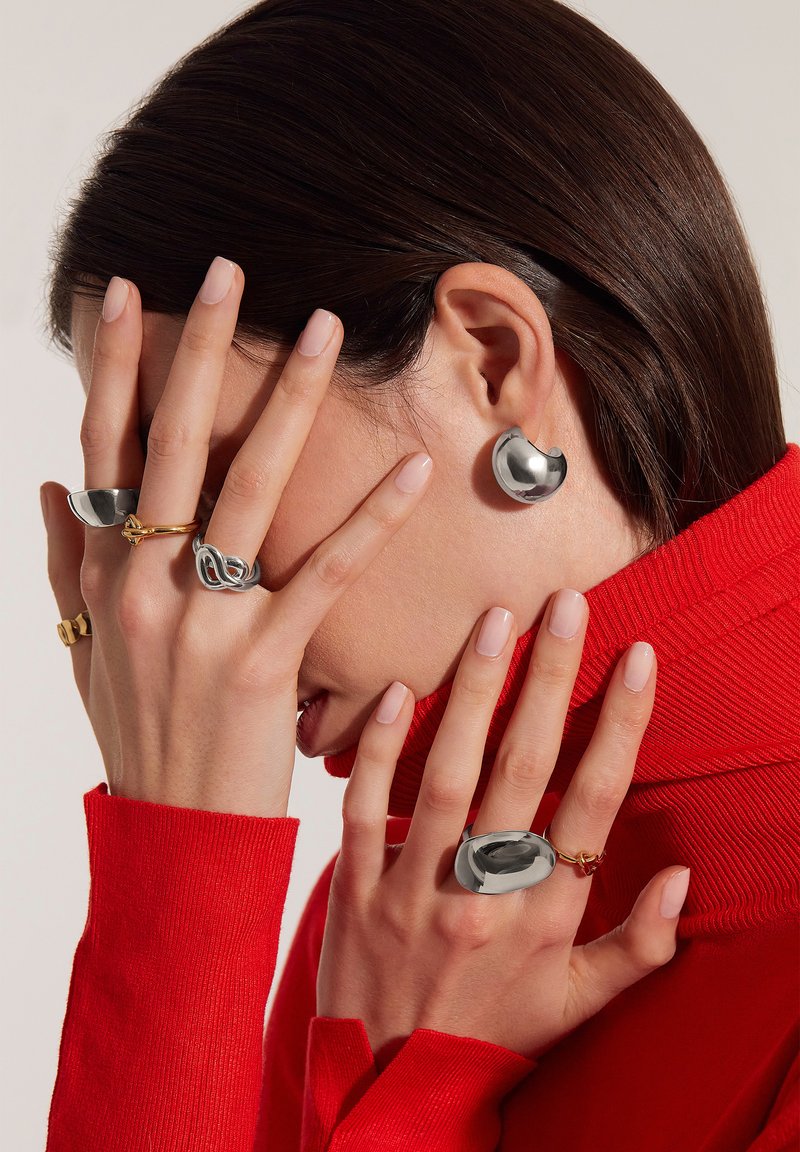 Orecchini d'argento con forme tondeggianti, grande anello d'argento, anello dorato intrecciato e dolcevita rossa a coste. Le unghie sono naturali.