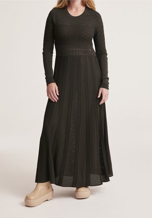 Vrouw die een lange mouw, donkergroene gebreide maxi-jurk met textuurpatronen draagt en beige plateauzolen, staand tegen een effen achtergrond.