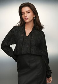 Blusa nera a maniche lunghe con motivo a foglia strutturato, scollo a V e dettaglio con coulisse. Abbinata a una gonna in pelle nera a vita alta.