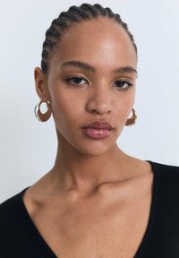 Gold hoop earrings, smooth texture, with a round shape and reflective finish. The subject wears a black V-neck top against a light background.