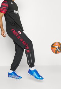 Svart atletisk klädsel med röda mönstrade detaljer på ärmarna och byxorna, kompletterad med blå fotbollsskor och en orange fotboll.