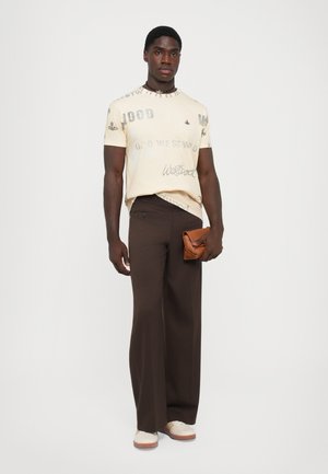 Man med en krämfärgad grafisk t-shirt, mörkbruna vida byxor, vita sneakers och hållande en ihopvikt brun läderclutch.