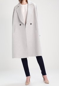 Cappotto oversize grigio chiaro con finitura texturizzata, due tasche frontali, design doppiopetto e bottoni marroni. Indossato con jeans scuri e tacchi.