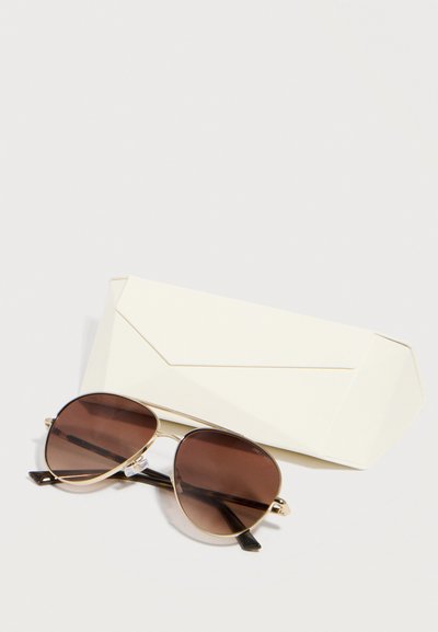 Ένα ζευγάρι αεροπόρων γυαλιά ηλίου με καφέ σκουρόχρωμους φακούς και χρυσά σκελετάκια που ακουμπούν πάνω σε μια κλειστή λευκή ορθογώνια θήκη πάνω σε λευκή επιφάνεια.