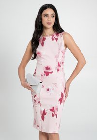 Figurbetontes pinkes Kleid mit floralem Muster in Rot und Weiß. Ärmelloses Design mit knielanger Schnittform. Hält eine metallisch silberne Clutch.