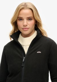Veste en polaire noire avec un col montant, fermeture éclair à l'avant et un petit logo sur la poitrine. Portée par-dessus un pull en maille côtelée crème.