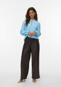 Donna che indossa una camicia a maniche lunghe azzurro chiaro, pantaloni a gamba larga marrone scuro e tacchi a punta con stampa rettile, in piedi davanti a uno sfondo bianco.