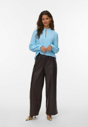 Mujer vistiendo una blusa de manga larga azul claro, pantalones de pierna ancha marrón oscuro y tacones de piel de serpiente puntiagudos, de pie contra un fondo blanco.