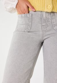 Pantaloni in denim grigio chiaro con tasche frontali. Presentano un design aderente con una texture liscia e dettagli di cucitura minimi. Abbinati a un maglione giallo.
