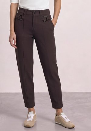 Femme portant un pantalon marron foncé sur mesure avec des détails de boucle argentée, associé à des baskets beiges et un haut blanc à manches courtes.