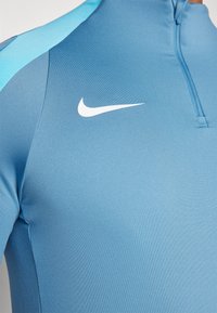 Világoskék sportos, félcipzáras felső sima anyagból, fehér Nike logóval a mellkason és kontrasztos kék vállrészekkel.