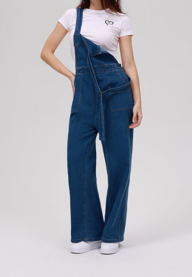 Femme portant une salopette en denim bleu à jambes larges avec une bretelle détachée, un t-shirt blanc avec un logo en forme de cœur, et des baskets blanches sur fond gris.