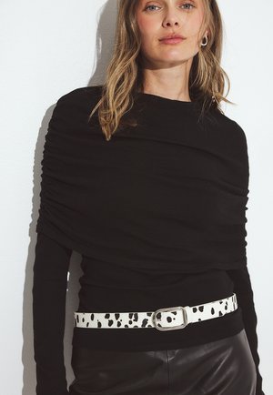 Žena oblečená v čiernom zberanom topu, s bielo-čiernym bodkovaným opaskom a čiernymi koženými nohavicami na pozadí bielej steny.